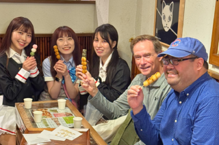 Five friends seated around a table, each holding a skewer of grilled dango or similar snacks and smiling at the camera.