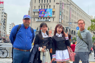 Four adults posing on a busy city street, two young women in matching black-and-white outfits flanking two men in casual clothes; urban shopping district backdrop.