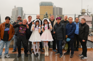 1 - SMILEYWALK Group of adults on a rooftop posing for a photo, with two women in white bunny-costumed dresses posing in front and a small Christmas tree behind them on a city skyline backdrop.
