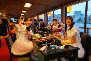 2 - SMILEYWALK Three young women in white feathered costumes dine at a long table on a boat restaurant, with city buildings and water visible outside the windows.