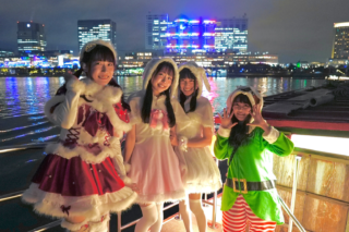4 - SMILEYWALK Four young women in festive cosplay pose on a lit waterfront boardwalk at night, with a city skyline and water reflections behind them.